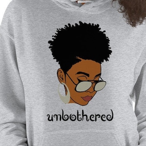 May include: Gray hooded sweatshirt with a black and brown illustration of a woman with short, curly hair wearing glasses and large hoop earrings. The text "unbothered" is printed below the illustration.