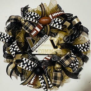 May include: A decorative wreath featuring black, gold, and white ribbons, with a football and a sign that says "Commodores" and "Vanderbilt". The wreath has a mix of patterned ribbons, including polka dots and plaid.