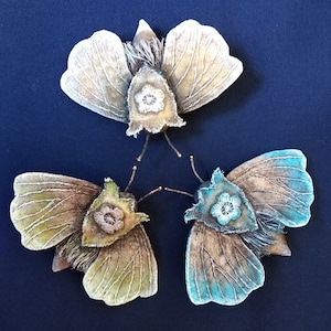 Puede incluir: Tres mariposas de tela bordadas con alas marrones, verdes y azules. Cada mariposa tiene un diseño de flor blanca en el centro de su cuerpo.