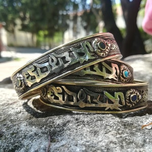 Puede incluir: Tres pulseras de plata con diseños intrincados y piedras preciosas de colores. Las pulseras están apiladas una encima de la otra y presentan escritura árabe.