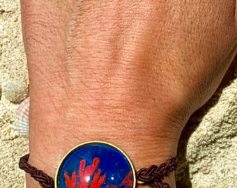 Puede incluir: Un brazalete trenzado marrón con un dije redondo de vidrio que presenta un diseño de coral rojo sobre un fondo azul.