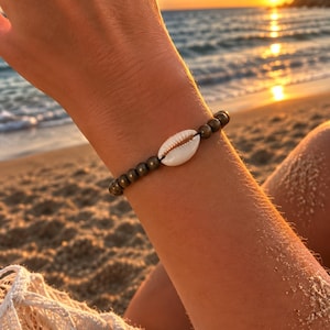 Puede incluir: Una pulsera de cuentas que presenta una sola concha de cauri blanca como punto focal central, flanqueada por una serie de pequeñas cuentas esféricas de color bronce metálico ensartadas en un cordón oscuro, creando un accesorio sencillo.