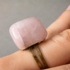 Puede incluir: Un anillo de cuarzo rosa con una piedra grande y rectangular. La piedra es de color rosa suave y tiene una superficie lisa y pulida. La banda del anillo es de metal de latón deslustrado.