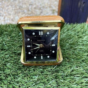 May include: A gold-toned alarm clock with a black face and white markings. The clock has a leather case and the words "Ingersoll-Waterbury" and "Alarm" are printed on the face.