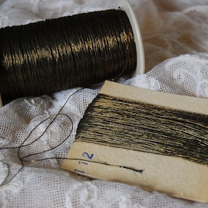 Può includere: Due rocchetti di filo metallico nero e oro. Uno è su una bobina di plastica bianca, l'altro su un piccolo cartoncino. Il filo è scuro con una lucentezza dorata metallica.