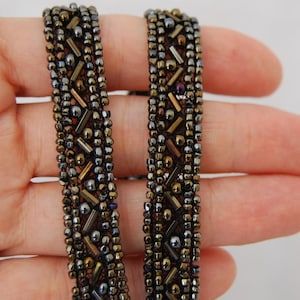 May include: A beaded trim featuring a repeating geometric pattern of metallic bronze, dark grey, and gunmetal seed beads and elongated bugle beads. The intricate embroidery is densely arranged on a dark fabric base, creating a textured band.