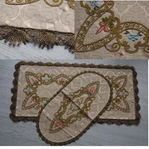 Könnte beinhalten: Beiges dekoratives Textil-Set mit aufwendiger Blumenstickerei in Rosa, Blau und Grün, umrandet mit Goldfaden. Das Set beinhaltet eine rechteckige Matte und ein ovales Stück, beide mit gemustertem Hintergrund und brauner Fransenborte.