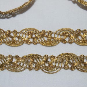Pode incluir: Close-up de acabamento decorativo dourado. O acabamento apresenta um design intrincado e vazado com padrões repetidos de laços e curvas. O acabamento é provavelmente usado para embelezar roupas ou artesanato.