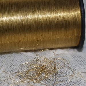 Może przedstawiać: Szpula złotej nici metalicznej z kilkoma nitkami rozwiniętymi i leżącymi na białej koronkowej powierzchni.
