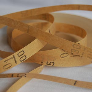 Puede incluir: Primer plano de un rollo de papel de lija de color beige con números negros impresos en la superficie. El papel de lija está enrollado y desenrollado, con los números 00, 50 y 30 visibles. El fondo es una superficie blanca lisa.