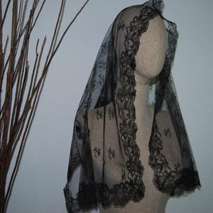 May include: A black lace veil featuring a delicate floral pattern throughout the fabric and a scalloped edge along the border. The sheer material drapes over a beige mannequin head and shoulders, showcasing the intricate needlework and design.