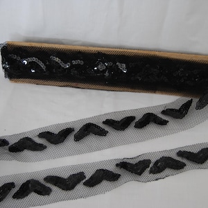 Peut inclure: Un rouleau de ruban en maille noire orné d'un motif répétitif de chevrons en sequins noirs. Le ruban est enroulé autour d'une bobine en bois rectangulaire, mettant en valeur la texture complexe et l'éclat réfléchissant des sequins sombres.