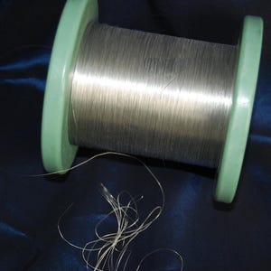 Pode incluir: Uma bobina de fio metálico prateado enrolado em torno de um núcleo de plástico verde claro. O fio tem um acabamento liso e reflexivo, enrolado firmemente em torno do cilindro central, com vários fios finos e soltos na base da bobina.