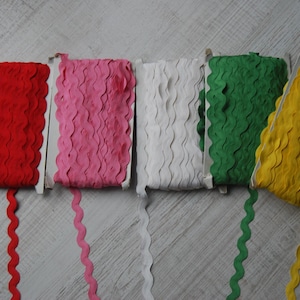 Puede incluir: Cinco carretes de cinta decorativa en rojo, rosa, blanco, verde y amarillo. Cada carrete es de un color diferente y tiene un borde festoneado. La cinta se muestra sobre una superficie de madera clara.