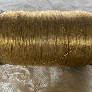 Könnte beinhalten: Eine Spule mit goldenem Metallic-Garn, das um eine Holzspule gewickelt ist. Das Garn ist glänzend und hat einen leichten Schimmer.