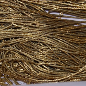 May include: A large collection of thin, flexible gold-colored snake chains intertwined in a dense pile. Each individual chain features a smooth, rounded, and tightly linked metallic texture that reflects light across the surface of the strands.