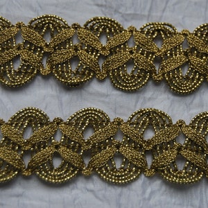 Pode incluir: Trimming metálico dourado com um design delicado e intrincado que apresenta motivos repetidos de folhas e círculos. O trimming é tecido e tem uma textura ligeiramente em relevo.