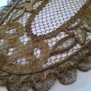 Op de afbeelding: Close-up van een vintage gouden kanten doily. De doily heeft ingewikkelde bloemen- en cirkelpatronen, met een delicaat, open geweven ontwerp. De randen zijn geschulpt, wat een vleugje elegantie aan het textiel toevoegt.