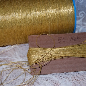 Puede incluir: Una bobina de hilo metálico dorado con una etiqueta que dice "50 metros". El hilo está enrollado alrededor de una bobina de cartón y está parcialmente desenrollado, mostrando la textura brillante y metálica.