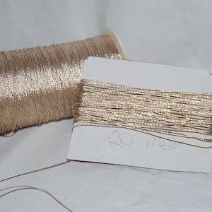 May include: A spool of metallic champagne-colored thread and a flat rectangular card wrapped with the same metallic thread. The card features the handwritten text 20 Meters, indicating the length of the thread wound around the cardboard base.