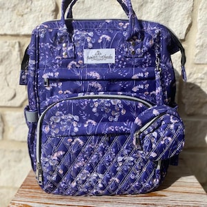 Peut inclure: Un sac à langer matelassé violet avec un imprimé floral et le texte "Sweet Cheeks Diaper Co.". Le sac a une poche zippée sur le devant et une petite pochette zippée attachée sur le côté.