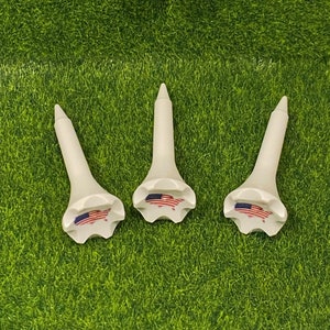 May include: Four white golf tees with a red, white, and blue American flag design. The tees are on a green artificial grass surface.
