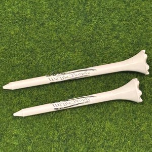 May include: Two white golf tees with the words "We the People" printed on them. The tees are on a green surface.