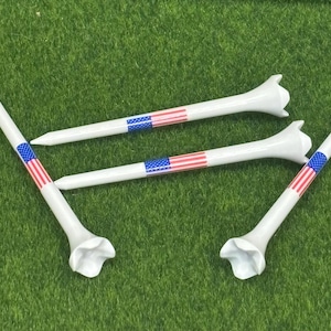May include: Four white golf tees with a design of the American flag on the shaft. The tees are arranged on a green surface, with the flag design featuring red and white stripes and blue stars.