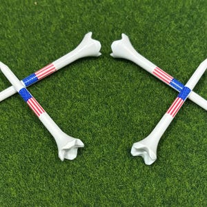 May include: Four white golf tees with a patriotic design. Each tee features a section decorated with the American flag, including red and white stripes and a blue field of stars. The tees are arranged on a green surface.