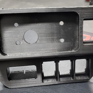 May include: A black plastic automotive dashboard component with rectangular and circular openings. The component has a textured surface and appears to be 3D-printed. It features a large rectangular opening and several smaller square openings.