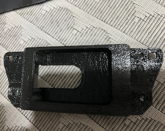 Peut inclure: Une pièce en plastique noir imprimée en 3D de forme rectangulaire avec une grande ouverture au centre. La pièce a une surface texturée et plusieurs petits trous autour des bords.