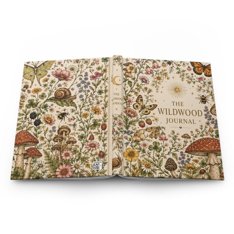 May include: Open journal with a beige cover featuring a whimsical floral and fauna design. The cover is adorned with butterflies, mushrooms, flowers, and the text "THE WILDWOOD JOURNAL". The journal's design has a vintage, nature-inspired aesthetic.