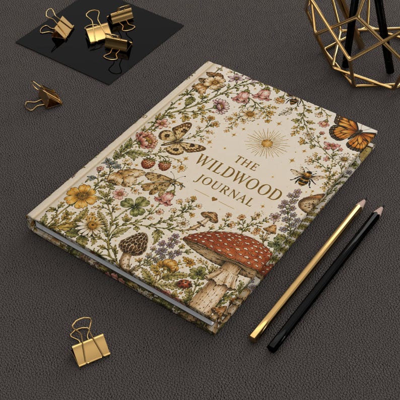 May include: A hardcover journal titled "The Wildwood Journal" with a floral and mushroom design. The cover features butterflies, bees, and a sun illustration. Gold and black pencils and gold binder clips are also present.