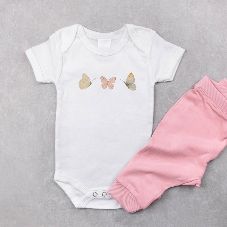 Puede incluir: Body de beb&eacute; blanco con tres ilustraciones de mariposas en tonos beige y rosa. Combinado con pantalones rosas para beb&eacute;. El conjunto se muestra sobre una superficie gris.