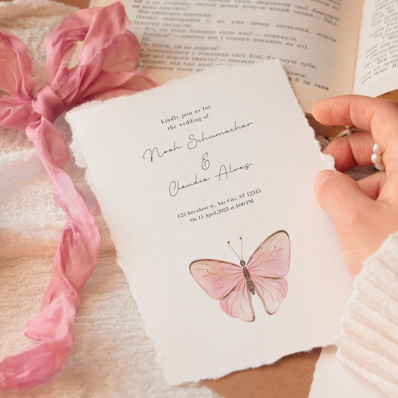 Puede incluir: Una invitaci&oacute;n de boda con una ilustraci&oacute;n de mariposa en acuarela. La invitaci&oacute;n presenta los nombres "Noah Schumacher & Claudia Alves" y los detalles del evento. Una cinta rosa y un libro est&aacute;n en el fondo.