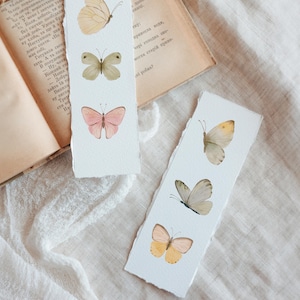 Puede incluir: Dos marcap&aacute;ginas de papel hechos a mano con ilustraciones de mariposas en acuarela. Un marcap&aacute;ginas presenta tres mariposas en tonos amarillos, verdes y rosas. El otro marcap&aacute;ginas tiene tres mariposas en tonos amarillos y grises.