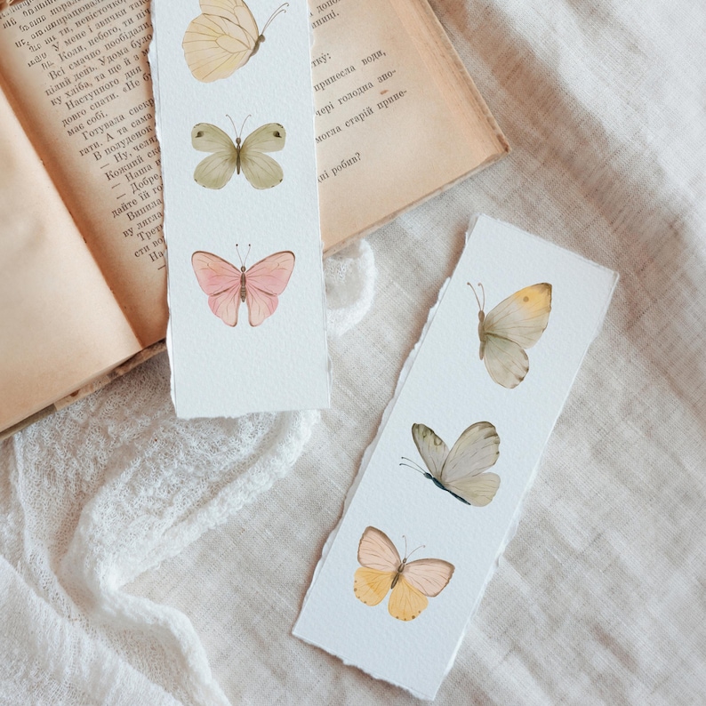 Puede incluir: Dos marcap&aacute;ginas de papel hechos a mano con ilustraciones de mariposas en acuarela. Un marcap&aacute;ginas presenta tres mariposas en tonos amarillos, verdes y rosas. El otro marcap&aacute;ginas tiene tres mariposas en tonos amarillos y grises.