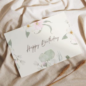 May include: A cream-coloured birthday card featuring the words "Happy Birthday" in script. The card is decorated with watercolour floral illustrations in white, pink, and green, with wheat sprigs in the foreground.