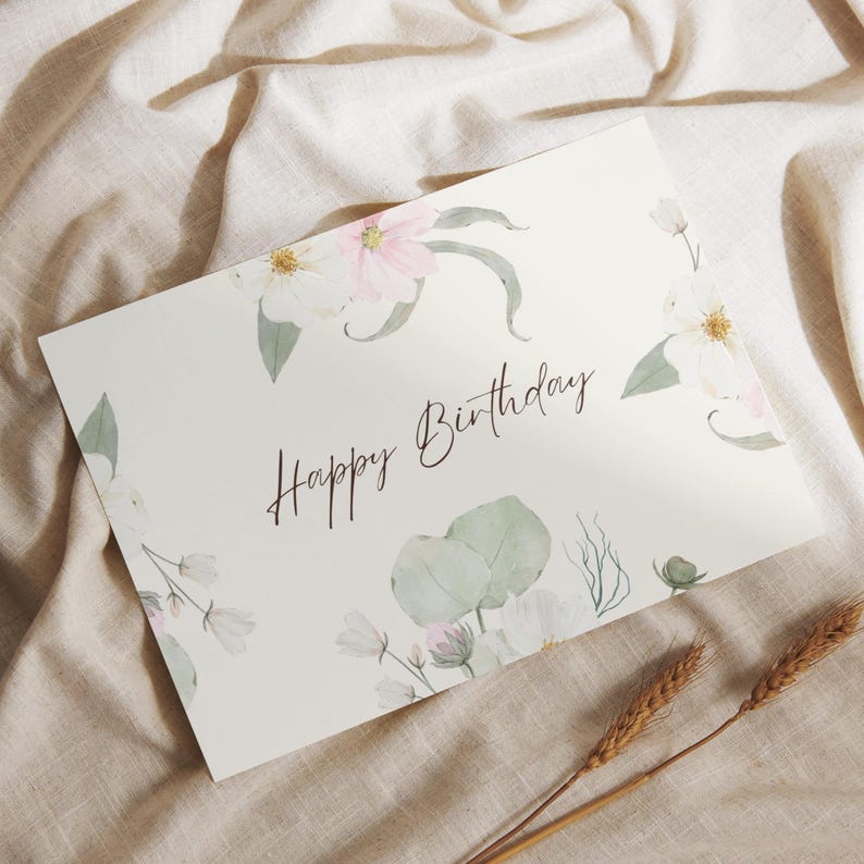 May include: A cream-coloured birthday card featuring the words "Happy Birthday" in script. The card is decorated with watercolour floral illustrations in white, pink, and green, with wheat sprigs in the foreground.