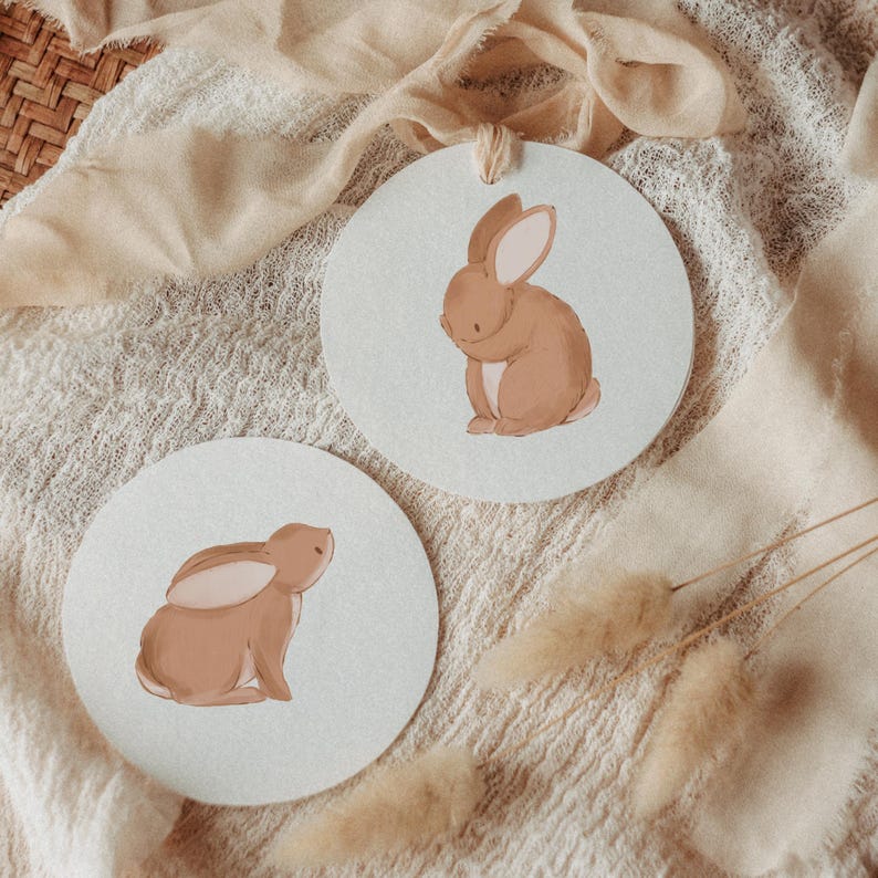 May include: Two round wooden ornaments with a white background and a brown bunny rabbit illustration on each. The bunny rabbits are in different poses.