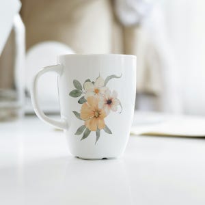 May include: White ceramic mug with a floral design. The design features yellow, peach, and white flowers and green leaves. The mug has a curved handle and a flared shape. The mug is on a white surface.