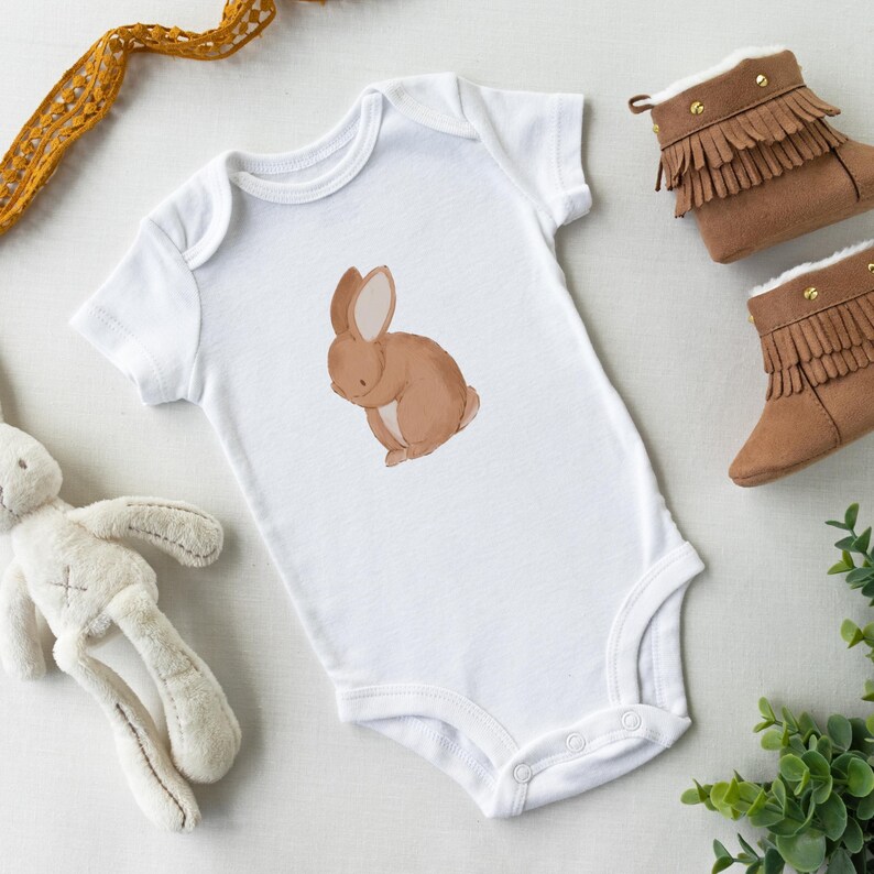 May include: A white baby onesie with a brown bunny rabbit illustration. The onesie has snaps at the bottom and is made of soft, comfortable fabric.