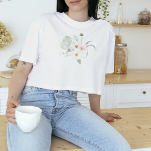 May include: White crop top with a watercolour floral design in soft pink, white, and green hues. The shirt is paired with light blue denim jeans. A white ceramic mug is held in the hand.