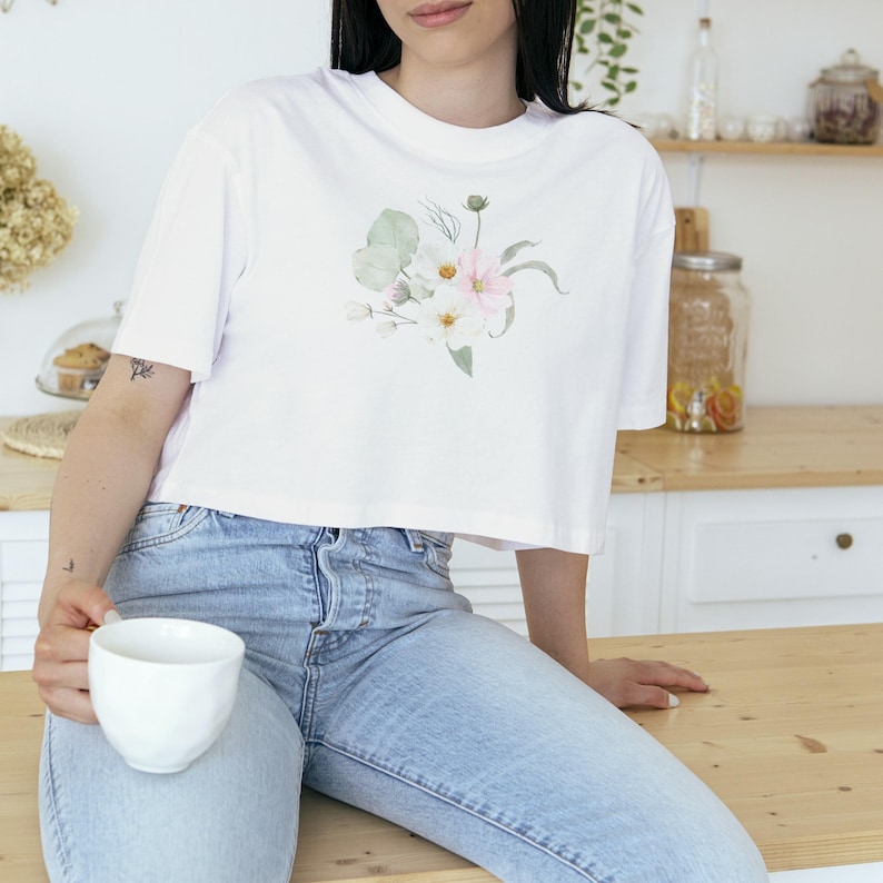 May include: White crop top with a watercolour floral design in soft pink, white, and green hues. The shirt is paired with light blue denim jeans. A white ceramic mug is held in the hand.