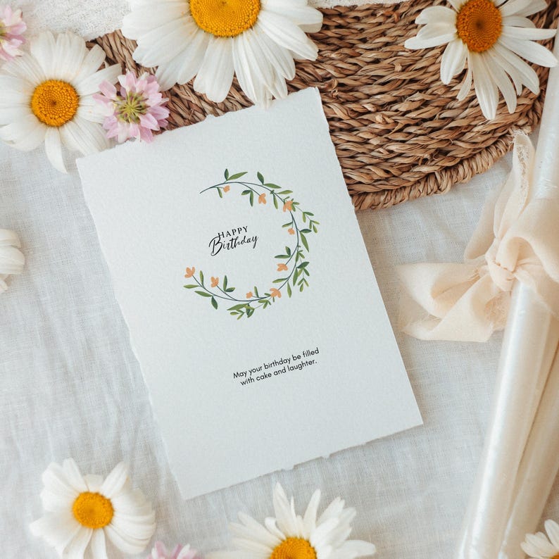 K&ouml;nnte beinhalten: Eine wei&szlig;e, rechteckige Gru&szlig;karte mit B&uuml;ttenrand, verziert mit einem gr&uuml;nen Blattkranz und kleinen orangefarbenen Bl&uuml;ten. Der Text lautet Happy Birthday und May your birthday be filled with cake and laughter in einer schwarzen Schrift.