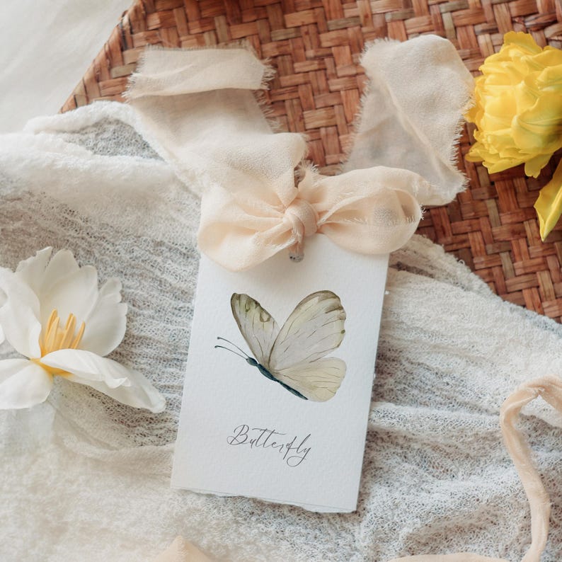 Puede incluir: Una tarjeta blanca con una ilustraci&oacute;n de mariposa en acuarela y la palabra "Butterfly" en escritura cursiva. La tarjeta est&aacute; atada con una cinta color melocot&oacute;n y colocada sobre una cesta tejida y tela blanca, con una flor blanca y una flor amarilla en el fondo.