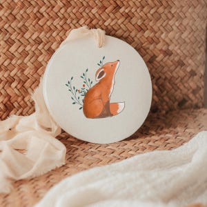 K&ouml;nnte beinhalten: Ein kreisf&ouml;rmiges, cremewei&szlig;es Keramikornament mit einer aufgedruckten Illustration eines sitzenden, nach oben blickenden orangefarbenen Fuchses, umgeben von zarten gr&uuml;nen Blattzweigen mit kleinen roten Beeren, aufgeh&auml;ngt an einem cremefarbenen Stoffband.