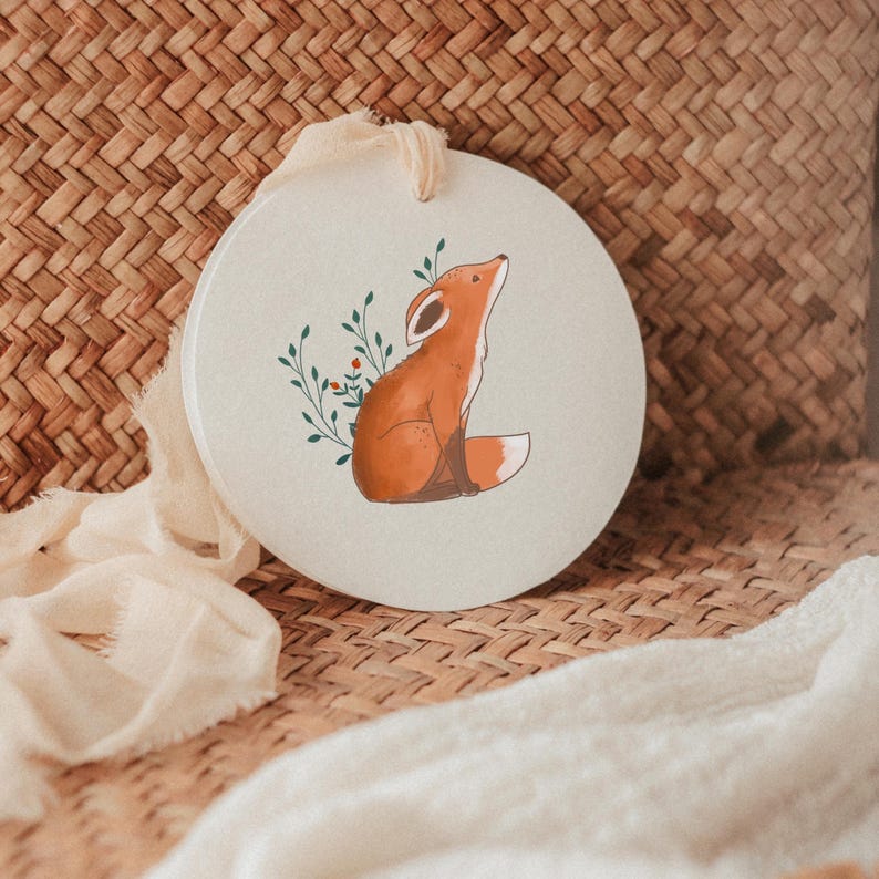 K&ouml;nnte beinhalten: Ein kreisf&ouml;rmiges, cremewei&szlig;es Keramikornament mit einer aufgedruckten Illustration eines sitzenden, nach oben blickenden orangefarbenen Fuchses, umgeben von zarten gr&uuml;nen Blattzweigen mit kleinen roten Beeren, aufgeh&auml;ngt an einem cremefarbenen Stoffband.