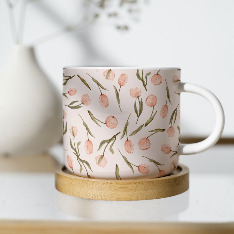 Puede incluir: Una taza de cer&aacute;mica blanca con un patr&oacute;n floral rosa y verde con tulipanes. La taza est&aacute; sobre un posavasos de madera.