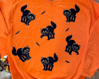 May include: Orange sweatshirt with a black cat design. The cats are in various poses and have green eyes. There are also small bat silhouettes on the sweatshirt.